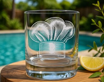 May include: A clear glass tumbler with a detailed engraving of a chef's hat in white and teal. The glass sits on a wooden surface, with a pool and greenery in the background. A slice of lemon is visible on the right.