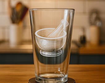 May include: A clear glass pint glass with a white etched design of a mortar and pestle. The glass sits on a round, dark brown coaster. The background is blurred, showing a kitchen setting with wooden surfaces.