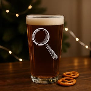 May include: A pint glass filled with dark beer and foam, featuring a white magnifying glass design. Two pretzels sit on a wooden surface in front of the glass. The background includes a blurred Christmas tree and string lights.