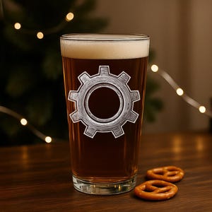 May include: A pint glass filled with beer and topped with foam. A white gear graphic is etched on the front of the glass. Two pretzels sit on a wooden surface in front of the glass. Fairy lights are in the background.