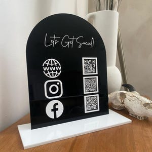 Könnte beinhalten: Ein schwarzes Social-Media-Schild aus Acryl mit weißem Sockel. Das Schild zeigt den Text "Lets Get Social!" und weiße Symbole für das Web, Instagram und Facebook sowie QR-Codes. Das Schild ist oben gewölbt.