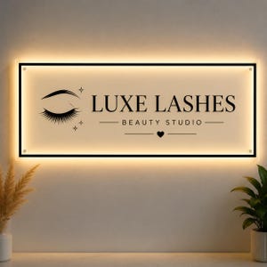 Könnte beinhalten: Ein rechteckiges Wandschild mit schwarzem Rahmen und warmer Hintergrundbeleuchtung. Das Schild zeigt den Text LUXE LASHES BEAUTY STUDIO in schwarzer Schrift, ergänzt durch eine Grafik eines geschlossenen Auges mit langen Wimpern.