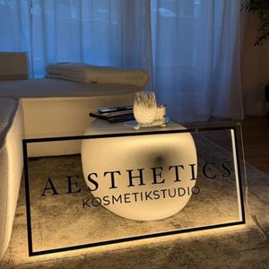Puede incluir: Letrero iluminado con las palabras "AESTHETICS KOSMETIKSTUDIO" en letras negras. El letrero es rectangular con un borde negro y está iluminado desde dentro. Una esfera brillante está detrás del letrero.