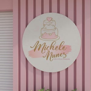 Könnte beinhalten: Ein rundes Schild mit einer rosa-weißen Kuchenillustration und dem Schriftzug "Michele Nunes" in goldener Schrift. Das Schild befindet sich vor einem rosa gestreiften Hintergrund, was auf eine Bäckerei hindeutet.