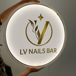 Könnte beinhalten: Rundes, beleuchtetes Schild mit weißer Oberfläche und goldenen Akzenten. Das Schild zeigt ein goldenes Logo mit einem Stern, Blättern und dem Buchstaben "V" in einem Kreis. Der Text "LV NAILS BAR" steht darunter.