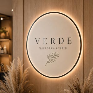 Könnte beinhalten: Ein kreisförmiges Wand-Schild mit schwarzem Rahmen und Hintergrundbeleuchtung. Das Schild zeigt den Text VERDE WELLNESS STUDIO in schwarzer Schrift, ergänzt durch eine einfache Strichzeichnung eines Blattzweigs unter dem Text.