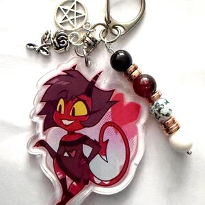 May include: A keychain with a cartoon demon character in red and black. It has a silver clasp, a pentagram charm, a rose charm, and a beaded accent with black, red, and white beads. The character has yellow eyes and a red tail.