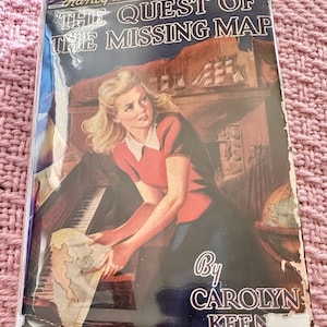 May include: A vintage book cover featuring a young woman in a red dress holding a map. The book title is "The Quest of the Missing Map" and the author is "Carolyn Keene". The book is part of the Nancy Drew Mystery Stories series.