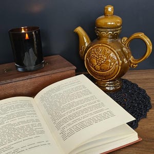 May include: A glossy brown ceramic teapot with a relief design of fruit in a basket, a lit candle in a dark glass jar, and an open hardcover book with white pages and black text. The items are arranged together on a flat wooden surface.