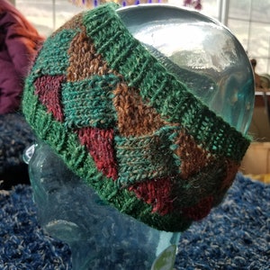 May include: A green, brown, and red knitted headband with a geometric pattern. The headband is made of wool and is soft and comfortable to wear.