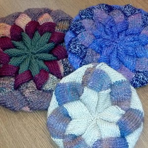 May include: Three circular knitted berets featuring a textured, woven-look pattern. One beret is primarily dark green and maroon, another is shades of blue and purple, and the third is a light cream color with blue and purple accents.