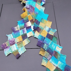 Puede incluir: Una bufanda de punto colorida con un diseño de patchwork en tonos de azul, verde, morado y dorado.