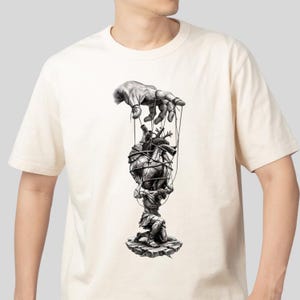 May include: Cream-colored t-shirt featuring a black and white illustration. The design depicts a hand manipulating a heart-shaped object with strings, held by a figure kneeling on a rock. The artwork has a detailed, artistic style.