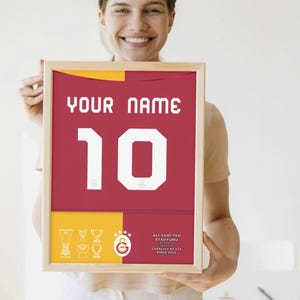 May include: A framed print of a red and yellow football shirt with the number 10 and the text "YOUR NAME". The shirt has a white star and the text "Ali Sami Yen Stadyumu" with the stadium's capacity and founding date.