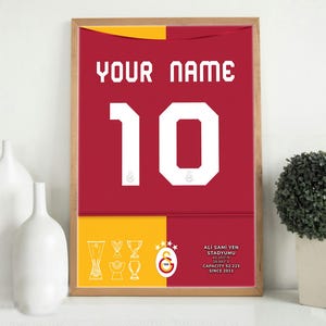 May include: A framed print of a red and yellow football shirt with the number 10 on the front. The shirt has the text "YOUR NAME" above the number and "ALI SAMI YEN STADIUMU" below the number. The text "CAPACITY 52,223 SINCE 2011" is also included.