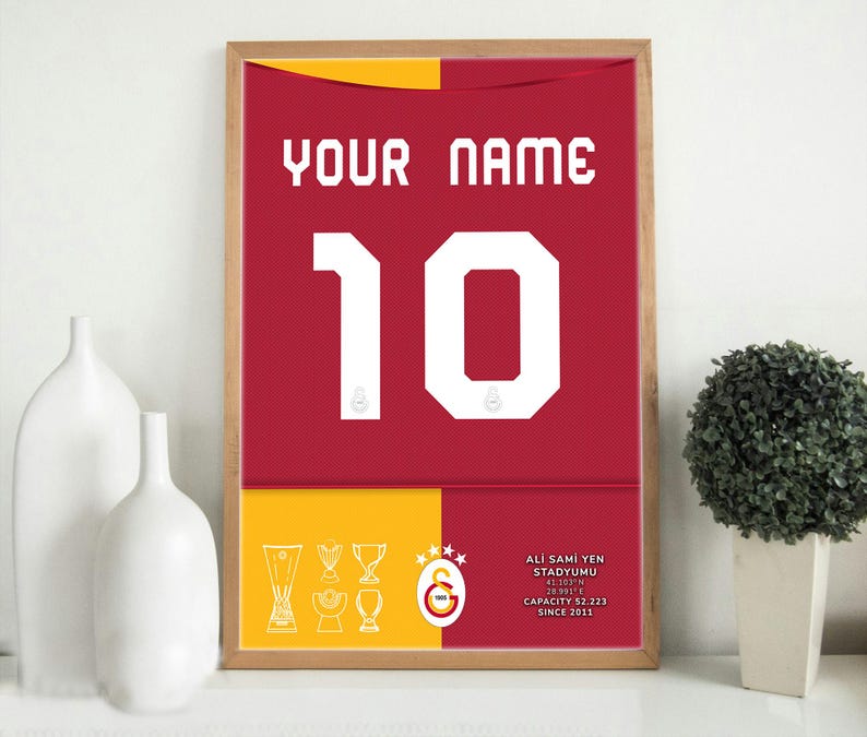 May include: A framed print of a red and yellow football shirt with the number 10 on the front. The shirt has the text "YOUR NAME" above the number and "ALI SAMI YEN STADIUMU" below the number. The text "CAPACITY 52,223 SINCE 2011" is also included.