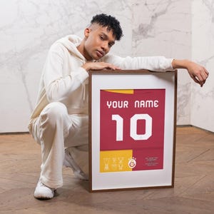 May include: A person is crouching in front of a framed jersey with the number 10 and the text "YOUR NAME". The jersey is red with yellow accents and a team logo.