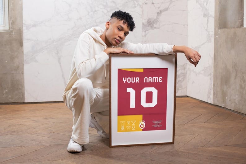May include: A person is crouching in front of a framed jersey with the number 10 and the text "YOUR NAME". The jersey is red with yellow accents and a team logo.