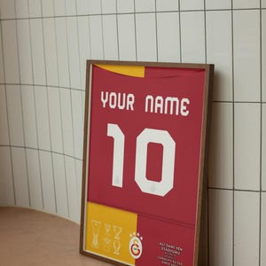 May include: A framed print of a red and yellow football shirt with the number 10 and the text "YOUR NAME". The shirt has a white star and crescent moon logo and the text "Ali Sami Yen Stadyumu" with the stadium's capacity and since date.