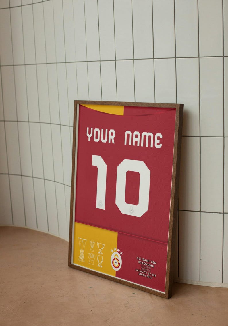 May include: A framed print of a red and yellow football shirt with the number 10 and the text "YOUR NAME". The shirt has a white star and crescent moon logo and the text "Ali Sami Yen Stadyumu" with the stadium's capacity and since date.