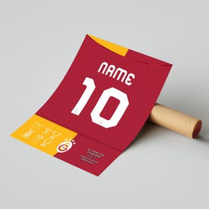 May include: A red and yellow jersey with the number 10 and the text "NAME" printed on it. The jersey is rolled up and has a brown cardboard tube next to it. The jersey has the text "Ali Sami Yen Stadyumu 1907-2011 Capacity 52,217 Since 2011" printed on the bottom.