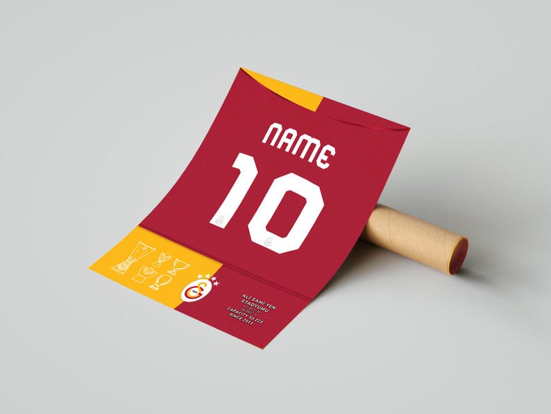 May include: A red and yellow jersey with the number 10 and the text "NAME" printed on it. The jersey is rolled up and has a brown cardboard tube next to it. The jersey has the text "Ali Sami Yen Stadyumu 1907-2011 Capacity 52,217 Since 2011" printed on the bottom.