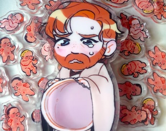 Puede incluir: Un llavero acrílico transparente con un personaje de dibujos animados con pelo rojo y barba, sosteniendo un objeto rosa. El llavero está rodeado de pequeños colgantes acrílicos de bebés en varias poses. El personaje tiene una expresión angustiada.