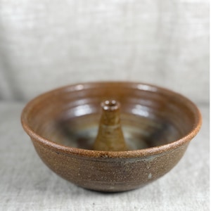 May include: A round, brown ceramic bowl with a central, tapered chimney-like pillar rising from the base. The exterior and interior surfaces feature a mottled, earthy glaze with subtle variations in tone and a textured, speckled finish.