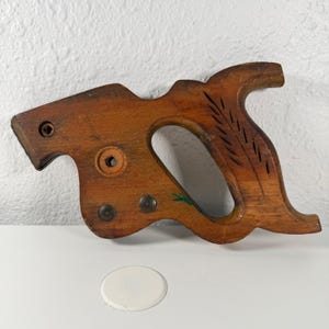 May include: A vintage wooden saw handle with a natural wood finish. The handle features a curved design with a large opening for gripping and decorative carved lines. It has metal rivets and a small green painted detail.