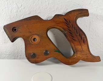 Könnte beinhalten: Ein Vintage-Sägegriff aus Holz mit natürlicher Holzoberfläche. Der Griff hat ein gebogenes Design mit einer großen Öffnung zum Greifen und dekorativen geschnitzten Linien. Er hat Metallnieten und ein kleines grün lackiertes Detail.