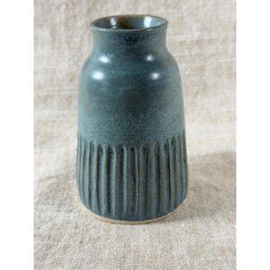 May include: A teal ceramic vase with a textured, ribbed design on the lower portion. The vase has a slightly flared top and a smooth, matte finish. The interior rim is a darker shade of green.
