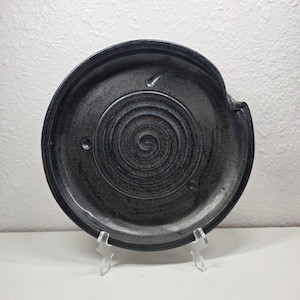 Puede incluir: Un plato de cerámica gris oscuro con un diseño en espiral en el centro. El plato tiene un borde ligeramente elevado y una pequeña extensión en forma de asa. Se muestra sobre un soporte transparente sobre un fondo blanco.