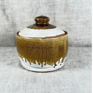 May include: A ceramic sugar bowl with a lid, featuring a white base and a brown glaze that drips downward from the top. The lid has a small, rounded knob at the center, and the overall finish shows a contrast between the dark brown and white.