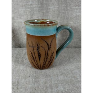Puede incluir: Una taza de cerámica con base marrón mate y un diseño grabado de tallos de trigo en color marrón oscuro. El borde superior y el asa curva tienen un acabado de esmalte turquesa brillante, con un acento marrón cálido en el borde interior.