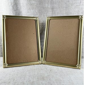 May include: Two rectangular picture frames with a metallic gold finish and decorative corner detailing. Each frame features a smooth, thin border surrounding a plain, light brown cardboard backing, presenting a clean and uniform appearance.