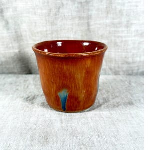 May include: A small, round ceramic cup featuring a glossy, warm orange-brown glaze with subtle vertical streaking. The interior is a deep, solid reddish-brown, and a single, distinct splash of bright blue glaze accents the lower exterior side.