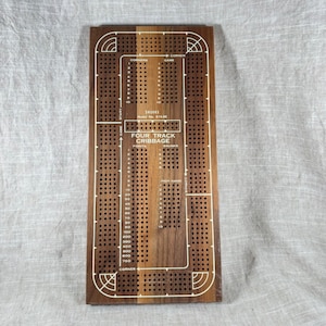 Può includere: Un tabellone di cribbage in legno con un design a quattro piste. Il tabellone è principalmente marrone con accenti di legno più chiari e segni bianchi. Il testo "FOUR TRACK CRIBBAGE" è visibile, insieme ad altre etichette e numeri relativi al gioco.