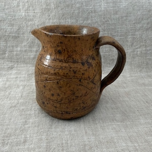 Può includere: Brocca in ceramica marrone fatta a mano, con manico curvo e beccuccio. La brocca ha una consistenza macchiettata e linee incise attorno al corpo. Il design rustico le conferisce un'estetica vintage e rustica.
