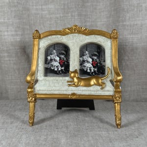 May include: A double photo frame with a gold-painted frame and legs, featuring an off-white textured center panel with a gold cat figure. The frame holds two black and white photos of children, each labeled with the text SIZE 2 x 3.5 INCHES.