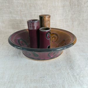 May include: A handmade ceramic flower frog with a deep burgundy and brown color scheme. The frog features a shallow dish with three cylindrical openings for holding stems. The dish has a black rim and decorative swirl patterns.
