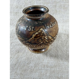 May include: A small, brown ceramic vase with a rounded body and a flared rim. The vase features an embossed design of a mountain and the text "Mt. St. Helens". The vase has a dark brown glaze.