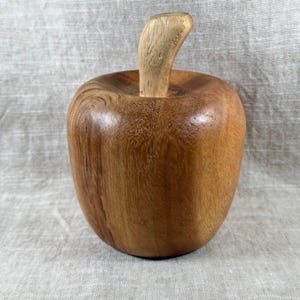 Pode incluir: Escultura de maçã de madeira com um caule castanho claro. A maçã é de uma cor castanha média quente, com grão de madeira visível. O caule é de um tom castanho mais claro. A escultura está sobre uma superfície de cor neutra.