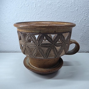 May include: A brown ceramic planter with a floral cut-out design around the middle. The planter has a matching saucer and a curved handle. The color is a mix of brown and tan.