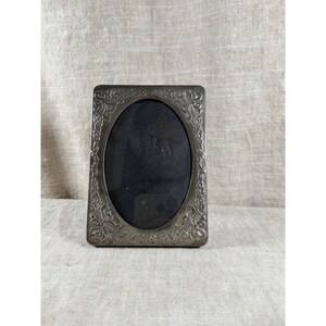 May include: A rectangular metal picture frame with a dark, tarnished silver finish. The frame features an oval-shaped central opening and is decorated with intricate, embossed floral and scrollwork patterns along the entire border.