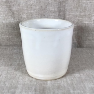 Peut inclure: Une tasse en céramique blanche avec une finition lisse et brillante et une forme cylindrique légèrement effilée. Le bord est arrondi et la base est plate, avec un vernis blanc cassé subtil qui recouvre toute la surface extérieure du récipient.