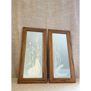 May include: Two rectangular wall mirrors with medium-toned wooden frames. Each mirror features a white etched design of underwater elements, including a seashell and seaweed on one, and an open clam shell with seaweed on the other.