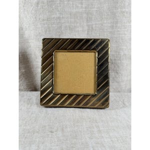 May include: A square-shaped picture frame with a metallic bronze finish. The frame features a prominent diagonal ribbed texture across its border, surrounding a plain, light tan square insert in the center. The design is uniform and symmetrical.