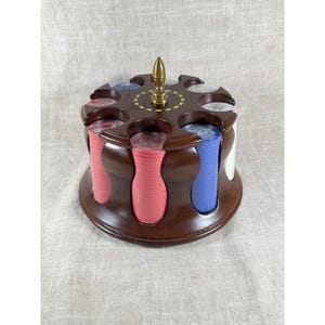 May include: A wooden poker chip carousel with a dark brown finish. The carousel holds stacks of red, blue, and white poker chips. A gold-colored decorative piece is on top of the carousel.