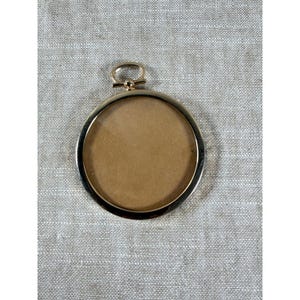 May include: A circular gold-toned metal frame featuring a smooth, polished finish and a clear glass center. The frame includes a small, rounded loop at the top for hanging or attachment purposes, presenting a simple and clean design structure.