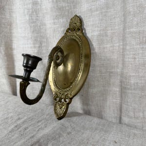 Puede incluir: Un candelabro de pared de latón con una placa ovalada que presenta motivos decorativos en forma de concha en la parte superior e inferior. Un brazo curvo y texturizado se extiende desde el centro, sosteniendo una pequeña copa cilíndrica.